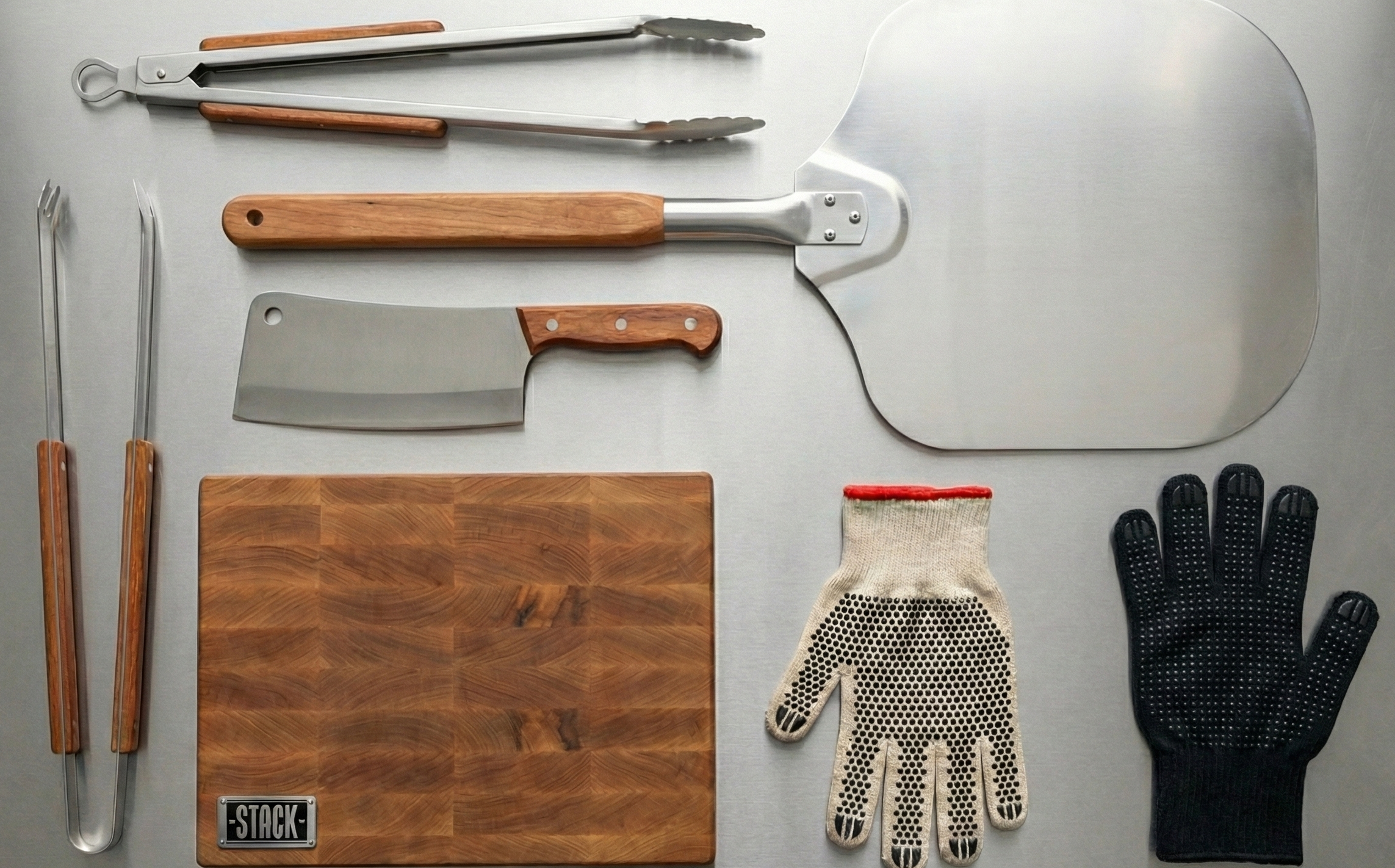Collection of barbecue tools including a cutting board, cleaver, spatula, tongs, and gloves on a gray background.
